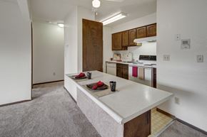 Anchorage apartment for rent - A practical kitchen here at The Continental showcasing dark wood cabinets, white appliances including a stove and dishwasher, laminate countertops with an extended breakfast bar, overhead lighting, and staged place settings that emphasize the open connection to the adjacent living area.