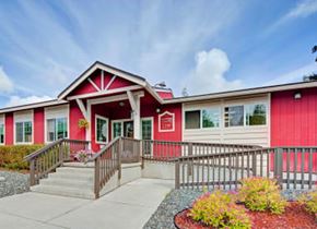 Anchorage apartment for rent - A bright community building exterior here at The Glen featuring a welcoming red-and-neutral facade with white trim, a central gabled entrance, and a fully accessible ramp and stairs leading to the front doors, surrounded by neatly landscaped beds, decorative gravel, and colorful shrubs under a partly cloudy blue sky.