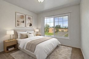 Anchorage apartment for rent - Bright bedroom here at The Glen featuring a neatly made bed with white bedding and neutral throw blanket, matching nightstands with lamps, soft carpet flooring, and a large window that brings in natural light and views of the community grounds.