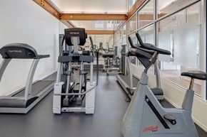Anchorage apartment for rent - A fitness center here at The Highlands Luxury, including a black and gray-colored treadmill, elliptical, and bike. The surrounding walls are open-concept glass walls with a mirror across the space, encompassed by soft-lighting.
