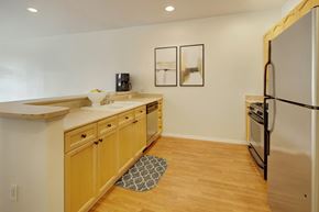 Anchorage apartment for rent - An open concept kitchen here at The Highlands Luxury, with light-colored wooden cabinets, a stainless steel fridge, oven, and dishwasher. A black coffee maker sits atop the cabinet. Includes a gray patterned kitchen mat on wooden floors. Kitchen overlooks into the living room.