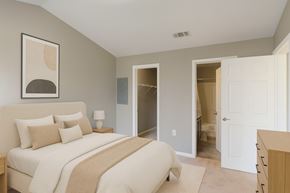 Bentonville apartment for rent - A calm bedroom here at Harbin Pointe with soft gray walls, plush carpet, and a cozy bed dressed in neutral bedding and layered pillows. A simple nightstand with a modern lamp sits beside the bed, with clean wall art above. The room includes an open closet and a doorway leading to the bathroom.