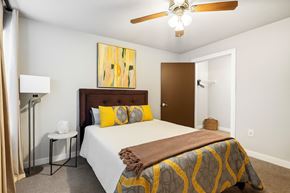 Rogers apartment for rent - A cozy, well-lit bedroom here at Palisades at Pleasant Crossing featuring a neatly made bed with neutral bedding and warm yellow accent pillows, a padded headboard, modern abstract wall art, soft carpet flooring, a ceiling fan with light, and an open closet that adds functional storage to the inviting space.