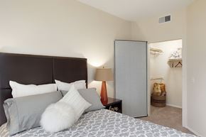 Phoenix apartment for rent - A cozy bedroom here at Acclaim featuring a plush bed with neutral bedding and layered accent pillows, a padded headboard, a bedside table with warm lamp lighting, soft beige carpeting, and an open walk-in closet with shelving and hanging space that adds everyday functionality.