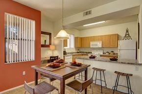 Phoenix apartment for rent - A welcoming dining and kitchen area here at Arezzo featuring a wooden dining table set beneath a pendant light, warm accent walls, an open breakfast bar with seating, light wood cabinetry, white appliances, and large windows with vertical blinds that bring in natural light.