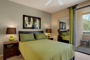 Phoenix apartment for rent - A stylish bedroom here at Aspire Pinnacle Park featuring a plush bed dressed in vibrant green bedding with coordinating patterned pillows and rich wood nightstands on either side. Matching table lamps add warm lighting, while abstract wall art and a mirrored closet reflect natural light from the sliding glass door leading to a private patio.