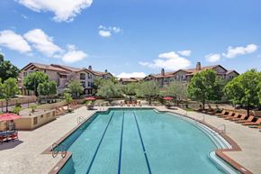 Anthem apartment for rent - A resort-style swimming pool here at Bela Rosa featuring a spacious deck with multiple lounge chairs and red umbrellas for shaded relaxation. Clear blue water with lap lanes creates a refreshing focal point, while surrounding trees and warm-toned apartment buildings provide a private, inviting atmosphere perfect for enjoying sunny Arizona days.