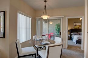 Chandler apartment for rent - A bright dining area here at Dobson Towne Center featuring plush carpet, a round dining table with seating for four, and a modern pendant light overhead. Large windows and a sliding glass door provide natural light and access to a private patio, while an open doorway leads to a spacious bedroom, creating a connected and comfortable layout.