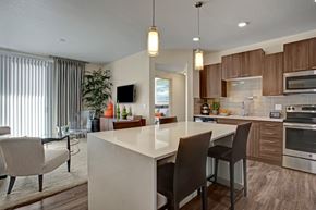 Phoenix apartment for rent - A contemporary open kitchen and living area here at Escape featuring rich wood-style cabinetry, stainless-steel appliances, and a sleek quartz island with bar seating under modern pendant lighting, complemented by wood-style flooring, soft carpeting in the living space, vertical blinds leading to a private balcony, and stylish furnishings that create a warm, inviting atmosphere.