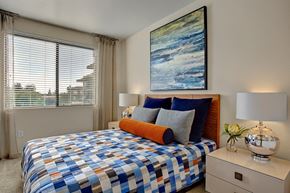 Phoenix apartment for rent - A stylish bedroom here at Escape featuring a warm wood headboard, layered blue and gray bedding with an accent bolster pillow, and matching nightstands topped with modern chrome lamps, while a large window with blinds and soft drapery fills the space with natural light and a bold abstract artwork adds a contemporary focal point above the bed.
