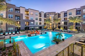 Gilbert apartment for rent - A resort-inspired pool courtyard here at The Flats at San Tan featuring a sparkling turquoise swimming pool with wide entry steps, surrounding lounge chairs with cushioned seating, illuminated palm trees, and a covered cabana with vibrant Adirondack chairs, all framed by contemporary apartment homes and warm evening lighting for a relaxing community retreat.