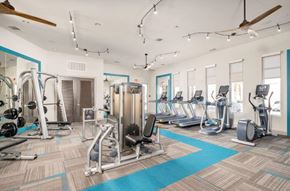 Gilbert apartment for rent - A spacious fitness center here at The Flats at San Tan featuring strength-training machines, free weights, and multiple cardio options including treadmills and elliptical machines, all arranged on patterned flooring with teal accents, large windows for natural light, ceiling fans, and modern lighting for an energizing workout environment.