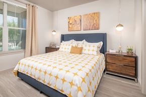 Gilbert apartment for rent - A serene and stylish bedroom here at The Flats at San Tan featuring a plush upholstered bed dressed in white bedding with golden patterned accents, complemented by matching nightstands, warm wood finishes, and modern pendant lighting, while a large window with soft curtains brings in natural light to create a bright and inviting retreat.
