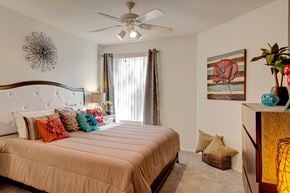 Phoenix apartment for rent - A cozy bedroom here at Hawthorne featuring a plush tufted headboard, soft neutral bedding accented with vibrant decorative pillows, and a ceiling fan with overhead lighting, complemented by a large window with vertical blinds and elegant drapery that invites natural light while maintaining privacy for a relaxing and comfortable retreat.