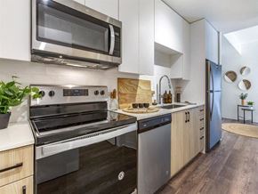 Phoenix apartment for rent - A sleek contemporary kitchen here at JOY on 4th featuring light wood cabinetry, white quartz countertops, and a modern tile backsplash. Stainless steel appliances, including a built-in microwave and full-size range, pair with a deep undermount sink and matte black hardware, while wood-style flooring flows into the open living area beyond.