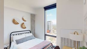 Phoenix apartment for rent - A bright and modern bedroom here at JOY on 4th featuring a sleek black metal bed frame with crisp white bedding and a soft accent throw, complemented by woven wall décor and a concrete accent column. Expansive floor-to-ceiling windows showcase sweeping city views while natural light enhances the clean lines, warm wood flooring, and contemporary finishes.