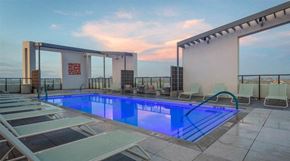Phoenix apartment for rent - A stunning rooftop pool deck here at JOY on 4th featuring a sleek rectangular swimming pool illuminated with vibrant blue lighting and surrounded by modern lounge chairs. Framed by clean architectural lines and glass railings, this elevated retreat offers sweeping city views, open sky sunsets, and stylish seating areas designed for relaxing, socializing, and soaking in the urban atmosphere.
