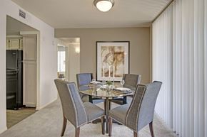 Phoenix apartment for rent - A warm welcoming dining area here at Mirador featuring plush carpet flooring and a round glass-top table surrounded by four upholstered chairs with nailhead trim. A large framed art piece anchors the space, while vertical blinds filter natural light and an open entryway leads toward the kitchen, creating an inviting setting for meals and gatherings.