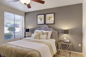 Phoenix apartment for rent - A cozy bedroom here at Mirador featuring a plush upholstered headboard, layered white and gold bedding, and two matching bedside tables with modern lamps. A large window with blinds invites natural light, while soft carpet flooring and neutral walls create a calm, comfortable retreat perfect for rest and relaxation.