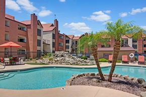 Phoenix apartment for rent - A resort-style pool area here at Motif featuring sparkling blue water, palm trees, and landscaped rock accents. Lounge chairs and shaded patio seating provide space to relax, while the surrounding three-story apartment buildings with balconies create a welcoming community atmosphere under a bright, sunny sky.