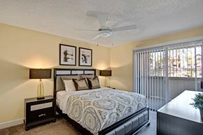 Phoenix apartment for rent - A cozy bedroom here at Motif featuring plush carpet, soft neutral walls, and a ceiling fan with light fixture. A large sliding glass door with vertical blinds opens to a private patio, bringing in natural light, while matching bedside tables and lamps create a balanced, inviting retreat.