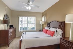 Phoenix apartment for rent - A comfortable bedroom here at Mozaic at Steele Park featuring plush carpet, neutral walls, and a ceiling fan with light. A large window with vertical blinds brings in natural light, while the spacious layout accommodates a full bedroom set including matching nightstands and a dresser for added storage.