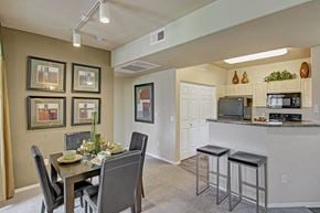 Peoria apartment for rent - A contemporary dining area here at Natura Villas featuring a dark wood table set for four with sleek upholstered chairs and decorative wall art. The space flows seamlessly into a bright kitchen with light cabinetry, a breakfast bar with seating, and modern appliances, creating an open and inviting layout ideal for everyday meals and entertaining.