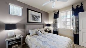 Peoria apartment for rent - A serene bedroom here at Natura Villas featuring a plush bed with patterned bedding and coordinating accent pillows, framed by soft neutral walls and a contrasting accent wall. Matching bedside tables with modern lamps add balance, while large windows with vertical blinds and layered curtains invite natural light, creating a calm and comfortable retreat.