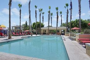 Peoria apartment for rent - A resort-style swimming pool here at Natura Villas surrounded by tall palm trees and expansive sun decks, featuring crystal-clear water, multiple lounge chairs with vibrant red cushions, and shaded seating areas under umbrellas. A covered pavilion offers additional gathering space, creating a relaxing outdoor retreat perfect for enjoying sunny Arizona days.