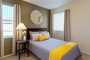 Mesa apartment for rent - A warm and inviting bedroom here at Pala Mesa featuring soft carpeting, a rich wood headboard, and neutral bedding accented with a vibrant yellow throw and decorative pillow. A bedside table with lamp adds cozy lighting, while two tall windows with vertical blinds and curtains fill the space with natural light.