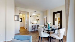 Mesa apartment for rent - A bright and welcoming dining area here at Pala Mesa featuring rich wood-style flooring, a round glass-top table set for four with upholstered cream chairs, and a bold turquoise vase centerpiece. The open pass-through to the kitchen includes a breakfast bar with stools, while neutral walls and a large framed mirror create an airy, spacious feel.