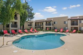Phoenix apartment for rent - A resort-style swimming pool here at Paradise Falls featuring a curved pool with clear blue water, surrounding concrete deck, and a row of red lounge chairs along a black metal fence, with mature trees and tan apartment buildings in the background creating a relaxing outdoor setting under open skies.