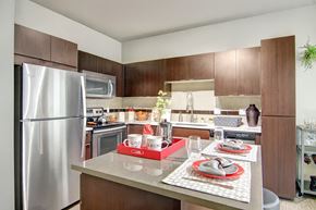 Phoenix apartment for rent - A modern kitchen here at Proxy 333 showcasing rich dark wood cabinetry, smooth light countertops, and a spacious island set with place settings, complemented by stainless steel appliances, a tiled backsplash, undercabinet lighting, and clean contemporary finishes that create a polished and inviting cooking and dining space.
