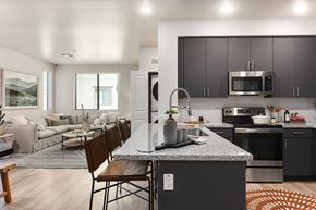 Mesa apartment for rent - A modern open living space here at Reflect at Dobson Ranch showcasing a contemporary kitchen with dark flat-panel cabinetry, a spacious island with bar seating, polished countertops, and sleek appliances, opening into a comfortable living room with a large neutral sectional, layered rugs, soft natural light, and warm wood-style flooring.