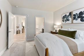 Mesa apartment for rent - A tranquil bedroom here at Reflect at Dobson Ranch showcasing a plush upholstered bed with crisp white bedding, a soft neutral throw, and layered accent pillows, paired with warm wood nightstands and modern table lamps, with framed botanical artwork above the headboard, plush carpeting underfoot, and open doorways leading to an en suite bathroom and hallway.