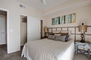 Tempe apartment for rent - A serene bedroom here at Rio Paradiso featuring a spacious bed dressed in neutral bedding with patterned accent pillows, matching bedside tables with decorative lamps, and soft carpet flooring. The room includes a ceiling fan, an open closet with shelving, and a private doorway leading to an adjoining bathroom for added comfort and convenience.