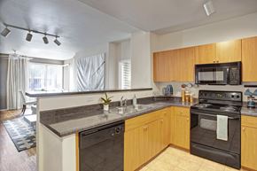 Tempe apartment for rent - A welcoming kitchen here at Rio Paradiso showcasing warm wood cabinets, dark countertops, a double sink with modern faucet, and matching black appliances. The layout includes a breakfast bar overlooking the living and dining area, track lighting overhead, and wood-style flooring that adds a cohesive, functional flow to the open-concept space.
