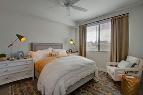 Phoenix apartment for rent - A serene bedroom here at Roosevelt Row featuring a plush upholstered bed dressed in layered neutral bedding, matching nightstands with modern accent lamps, a cozy armchair with a small side table, patterned area rug over wood-style flooring, a ceiling fan, and a large window with blinds and curtains allowing soft natural light to fill the space.
