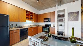 Phoenix apartment for rent - A contemporary kitchen here at Skyline Lofts showcasing warm wood cabinetry with open shelving, dark appliances, and sleek countertops, paired with an inset pantry nook and industrial-style concrete ceilings, while a glass dining table with modern place settings and decorative accents completes the open, loft-inspired layout.