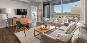 Tempe apartment for rent - A bright living room here at Skywater at Tempe Town Lake featuring a neutral sofa with textured throw pillows, two coordinating armchairs, and a warm wood coffee table set on a plush area rug over wood-style flooring, accented by a sculptural chair, modern floor lamp, ceiling fan, and large windows that bring in abundant natural light and views of the surrounding landscape.