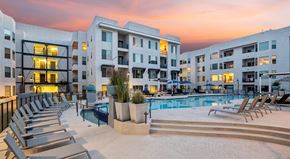Tempe apartment for rent - A resort-style courtyard pool here at Skywater at Tempe Town Lake showcasing a large swimming pool with a raised spa, tiered steps, and surrounding lounge chairs, framed by modern mid-rise apartment buildings, landscaped planters, and ambient lighting under a colorful sunset sky that creates a relaxing community atmosphere.