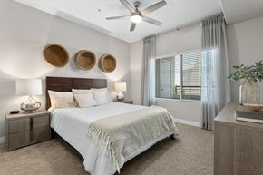 Tempe apartment for rent - A bright bedroom here at Skywater at Tempe Town Lake featuring a wooden bed frame with crisp white bedding and textured throw, matching nightstands with table lamps, woven wall décor above the headboard, soft carpet flooring, a ceiling fan, and a large window with sheer curtains that bring in natural light.