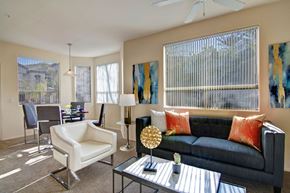 Gilbert apartment for rent - A welcoming living room here at Sonoma Landing featuring a dark upholstered sofa accented with warm orange pillows, a sleek glass coffee table with modern décor, and a white accent chair, while large windows with vertical blinds fill the space with natural light and connect to a dining area with a contemporary table, chairs, and pendant lighting.