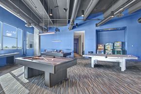 Phoenix apartment for rent - A vibrant resident game room here at Trend at 51 featuring multiple pool tables with overhead pendant lighting, a ping pong table, classic arcade machines along a bold blue accent wall, exposed industrial-style ceilings with visible ductwork, patterned carpeting, and large windows that bring in natural light and views of the surrounding area.