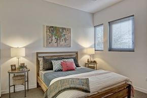 Phoenix apartment for rent - A serene bedroom here at Trend at 51 featuring a wood-frame bed dressed in layered neutral and blue bedding with accent pillows, matching bedside tables topped with modern lamps, soft carpet flooring, and abstract wall art above the headboard, while tall windows with blinds bring in natural light and create a calm, inviting atmosphere.