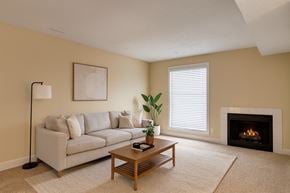 Calgary apartment for rent - A cozy living room here at 3600 Brentwood with soft beige carpet, a light gray sofa, a wooden coffee table, and a warm fireplace, accented by a bright window with white blinds and simple modern decor.