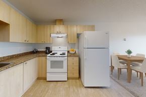 Wetaskiwin apartment for rent - A spacious kitchen here at Birch Terrace with light wood cabinets, granite-style countertops, and white appliances including a stove and refrigerator. A dining area with a simple table and upholstered chairs sits nearby, creating a bright, open layout.