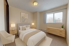 Edmonton apartment for rent - A cozy bedroom here at Broadmoor with soft carpet, a neatly made bed with neutral bedding, and warm bedside lighting. A large window brings in natural light, while a light wood dresser, accent chair, and simple wall art create a calm, inviting feel.
