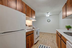 Edmonton apartment for rent - A spacious kitchen and dining area here at Cambridge West featuring rich wood cabinets, white appliances, and a clean countertop with a built-in sink. The open floor space with wood-style flooring, bright stove lighting, and a ceiling fan creates a simple, functional layout with room for a dining table.