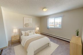 Edmonton apartment for rent - A bright bedroom here at Cambridge West with a soft upholstered bed, crisp white bedding, and matching nightstands with warm table lamps. A large window brings in natural light, while neutral carpeting, simple wall art, and a light wood dresser with a small plant create a calm, comfortable space.